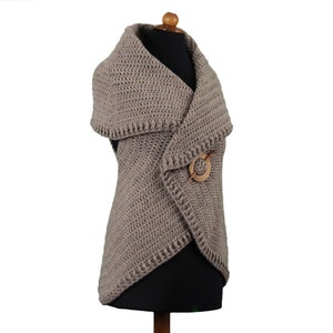 May include: A taupe-colored, crocheted vest with a shawl collar and a wooden pin closure. The vest is displayed on a mannequin and has a textured stitch pattern. The wooden pin is circular with a pin through the center.