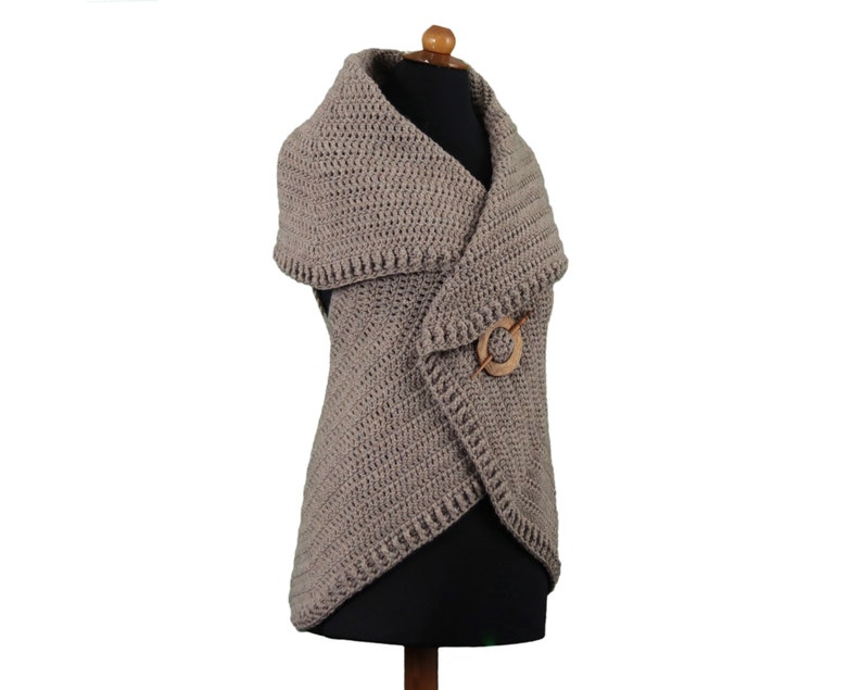 May include: A taupe-colored, crocheted vest with a shawl collar and a wooden pin closure. The vest is displayed on a mannequin and has a textured stitch pattern. The wooden pin is circular with a pin through the center.