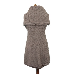 May include: A taupe-colored, handmade crochet vest with a wide, folded collar. The vest is sleeveless and has a slightly flared shape, with a detailed crochet stitch pattern. The garment is displayed on a wooden mannequin, showcasing its texture and design.