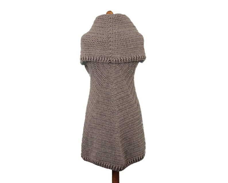 May include: A taupe-colored, handmade crochet vest with a wide, folded collar. The vest is sleeveless and has a slightly flared shape, with a detailed crochet stitch pattern. The garment is displayed on a wooden mannequin, showcasing its texture and design.