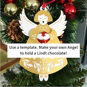 May include: A gold and white die-cut paper angel ornament featuring a central circular cutout designed to hold a round chocolate. The angel has stylized wings and a floral pattern at the base. Text reads: Use a template. Make your own Angel to hold a Lindt chocolate!