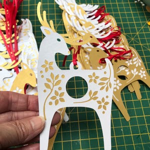 May include: A stack of white and gold paper reindeer ornaments with a hole in the center for hanging. The reindeer have a floral design and are cut out with a laser.