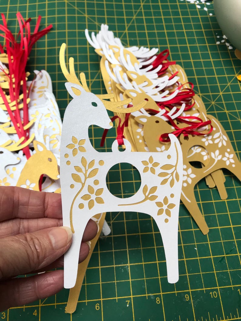 May include: A stack of white and gold paper reindeer ornaments with a hole in the center for hanging. The reindeer have a floral design and are cut out with a laser.
