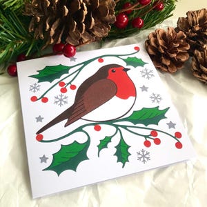 Modèle Robin sur houx. Carte de voeux de Noël. Couleurs traditionnelles. Blanc à l'intérieur. Impression d'oeuvres d'art originales découpées dans du papier. 148 x 148 mm.