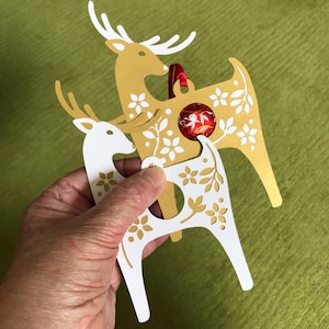 May include: Two gold reindeer ornaments with white floral designs. The reindeer are stacked on top of each other. The top reindeer has a red ribbon and a red and white candy ornament hanging from it.