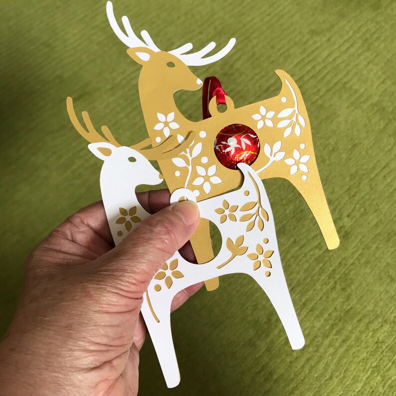 May include: Two gold reindeer ornaments with white floral designs. The reindeer are stacked on top of each other. The top reindeer has a red ribbon and a red and white candy ornament hanging from it.