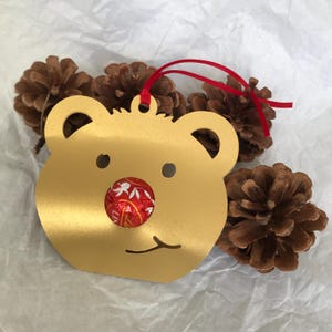 May include: Gold-colored metal bear ornament with a red and white candy nose. The ornament has a red ribbon attached for hanging.