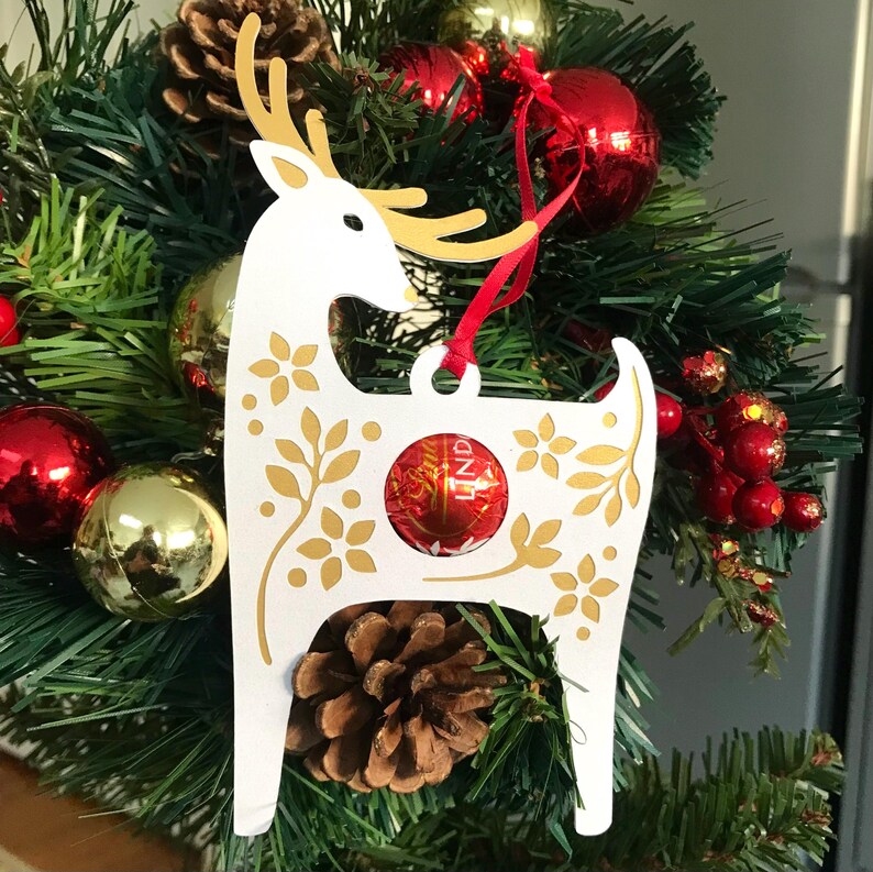 May include: White reindeer Christmas ornament with gold floral details and a red and gold Lindt chocolate inside. The ornament is hanging on a green Christmas tree.