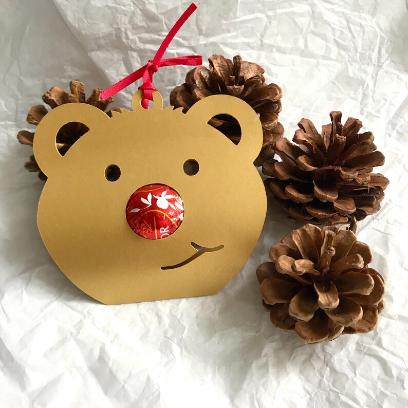 May include: A gold cardboard bear-shaped ornament with a red and white candy in the nose. The ornament is hanging from a red ribbon.