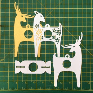 May include: Three paper cut-out reindeer ornaments, two white and one yellow, with floral designs. The reindeer are standing on a green cutting mat with a ruler.