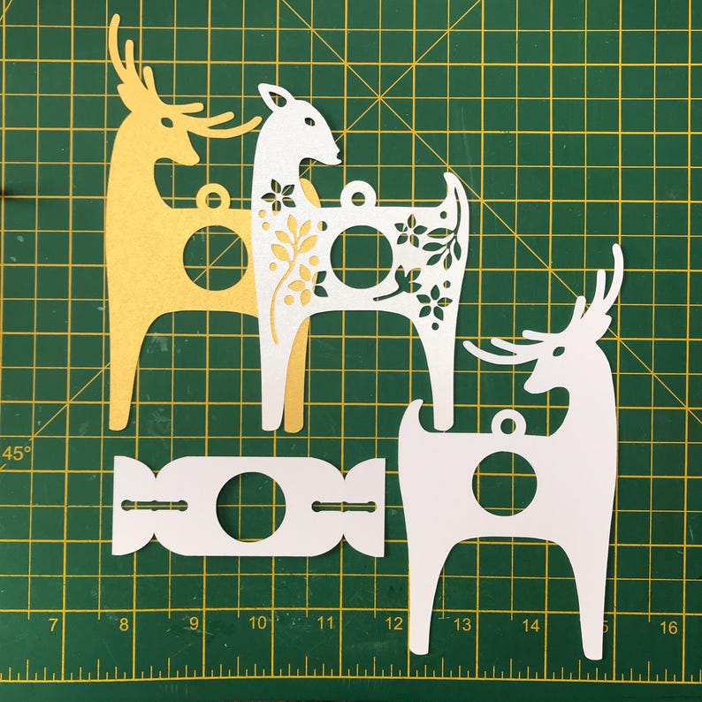 May include: Three paper cut-out reindeer ornaments, two white and one yellow, with floral designs. The reindeer are standing on a green cutting mat with a ruler.