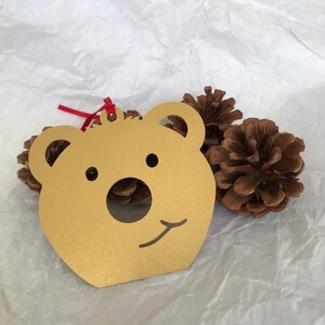May include: Gold-colored metal bear ornament with a red ribbon. The bear has a simple, cartoon-like face with a small nose and a smile.