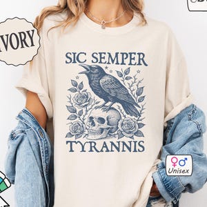May include: Ivory-colored unisex t-shirt with a graphic featuring a crow perched on a skull, surrounded by roses, and the text "SIC SEMPER TYRANNIS." The design is in shades of blue. The shirt is customizable.