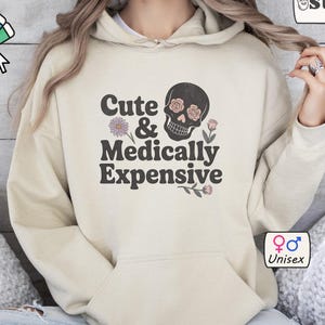 May include: A beige unisex hoodie with a front pocket and a drawstring hood. The graphic on the front says "Cute & Medically Expensive" with a skull and floral design. The hoodie is made of a soft material.