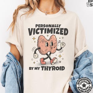 May include: A beige t-shirt featuring a cartoon thyroid gland with arms, legs, and bandages. The text reads "PERSONALLY VICTIMIZED BY MY THYROID" in bold black letters. The graphic includes small star and musical note accents surrounding the character.