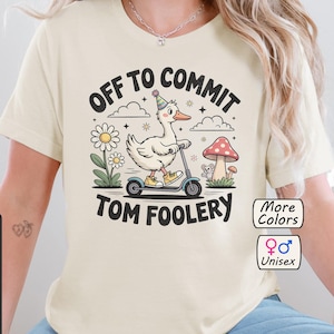 Könnte beinhalten: Cremefarbenes Unisex-T-Shirt mit dem Text "OFF TO COMMIT TOM FOOLERY". Die Grafik zeigt eine Ente auf einem Roller, die einen Partyhut trägt und von Blumen und einem Pilz umgeben ist. Das Shirt ist von Bella+Canvas.