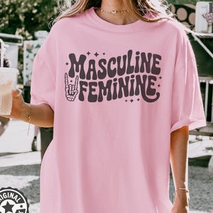 May include: A light pink t-shirt with the words "MASCULINE FEMININE" in a groovy font. The design includes a hand making a rock and roll sign with a skeleton hand. The shirt is a relaxed fit.