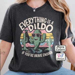 Könnte beinhalten: Ein dunkelgraues T-Shirt mit einem Grafikdruck eines Cartoon-Kaktus mit Sonnenbrille und dem Text "Everything is a Dildo If You're Brave Enough". Die Grafik ist vor einem Sonnenuntergangs-Hintergrund mit einem UFO am Himmel platziert. Das T-Shirt ist unisex und in verschiedenen Farben erhältlich.