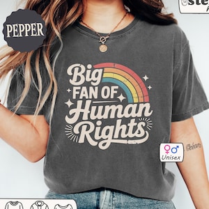 May include: Pepper-colored unisex t-shirt with the text "Big Fan of Human Rights" in a retro font, featuring a rainbow design. The shirt is from Comfort Colors and has a relaxed fit. The shirt is a unisex style.
