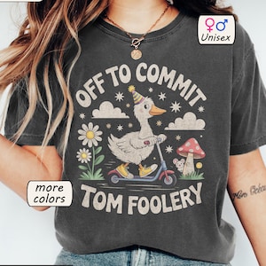 May include: Dark grey unisex t-shirt featuring a cartoon duck on a scooter, wearing a party hat, and the text "OFF TO COMMIT TOM FOOLERY." The shirt also has the text "more colours" and "Unisex."