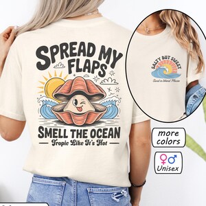 May include: Cream-colored unisex t-shirt with a graphic of a winking clam shell, sun, and waves. Text reads "SPREAD MY FLAPS" and "SMELL THE OCEAN - Tropic Like It's Hot." Additional text: "more colors" and "Unisex."