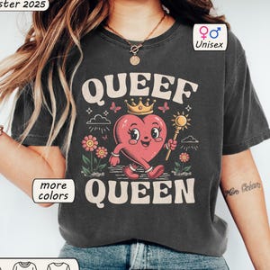 Pode incluir: Uma camiseta cinza escura com um coração de desenho animado usando uma coroa e segurando um cetro. O texto "Queef Queen" está impresso na camiseta.