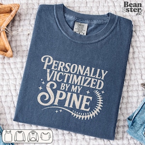 May include: A blue Comfort Colors t-shirt with the text "Personally Victimized by my Spine" in a white, decorative font. The shirt has a crew neck and a tag at the neckline. The shirt is folded neatly on a white surface.