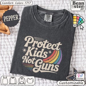 May include: A dark gray unisex t-shirt with the text "Protect Kids Not Guns" in a retro font, accompanied by a rainbow graphic. The shirt is customizable and made of Comfort Colors material.