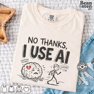 May include: A cream-coloured t-shirt featuring the black text "NO THANKS, I USE AI". Below the text is a graphic of a sad, crying brain with a red broken heart and a stick figure walking away with a speech bubble that says "LATER!".