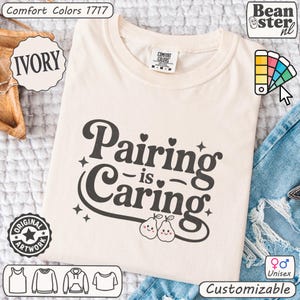 Peut inclure: T-shirt ivoire avec l'inscription "Pairing is Caring" dans une police fantaisiste, ornée de deux illustrations de poires souriantes. Le t-shirt a un col rond et des manches courtes. Personnalisable et unisexe.