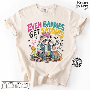 May include: Cream-colored t-shirt featuring a distressed raccoon with a pink mohawk, denim vest, and the text "EVEN BADDIES GET SADDIES NO FUTURE." The raccoon is sitting on a brick wall with a skateboard and spray paint.