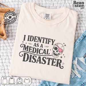 May include: Cream-colored t-shirt with the text "I IDENTIFY AS A MEDICAL DISASTER" and a skull graphic wearing an eye patch. The shirt has a crew neck and short sleeves. The shirt is folded on a white surface.