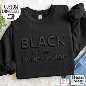 Op de afbeelding: Zwart sweatshirt met de geborduurde tekst "BLACK is my happy color". De sweatshirt heeft een ronde hals en lange mouwen. De afbeelding bevat ook een "Custom Embroidery" logo en een "Unisex" symbool.