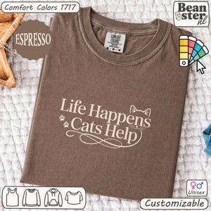 May include: Espresso-colored unisex t-shirt with the text "Life Happens Cats Help" in white. The shirt is from Comfort Colors and is customizable. The design includes a cat silhouette and paw prints.