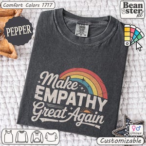 May include: A dark gray unisex t-shirt with the text "Make EMPATHY Great Again" in a vintage style with a rainbow and sun graphic. The shirt is customizable and made of Comfort Colors material.