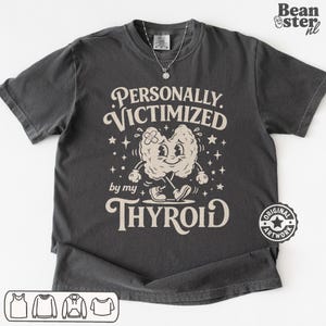 May include: Dark gray t-shirt with the text "PERSONALLY VICTIMIZED by my THYROID" and a cartoon thyroid graphic. The graphic is beige with a bandage and walking legs. A silver necklace is visible.
