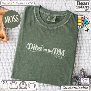 May include: Moss green unisex t-shirt with the text "Dibs on the DM" in white. The shirt is from Comfort Colors and is customizable. The design includes dice graphics, and the shirt is available in various styles.