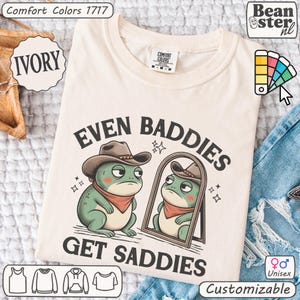 May include: Ivory-colored t-shirt with the text "EVEN BADDIES GET SADDIES" and a cartoon frog wearing a cowboy hat and a red bandana. The shirt is customizable and unisex.