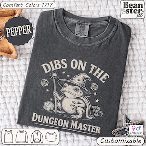 May include: Charcoal grey unisex t-shirt featuring the text "DIBS ON THE DUNGEON MASTER" and a frog in a wizard hat. The shirt is made of Comfort Colors 1717 material and is customizable. The t-shirt is a unisex design.