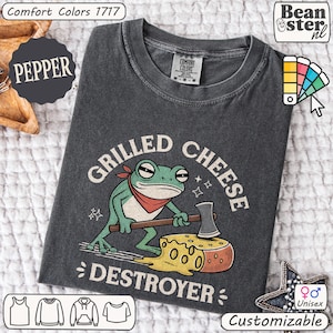 May include: Dark grey unisex t-shirt featuring a frog graphic with an axe and a cheese wheel. Text reads "Grilled Cheese Destroyer." The shirt is customizable and made of Comfort Colors material.
