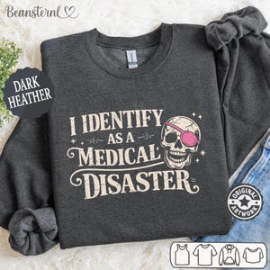 Może przedstawiać: Ciemnoszara bluza z napisem "I IDENTIFY AS A MEDICAL DISASTER" i czaszką z różową opaską na oku. Bluza z okrągłym dekoltem ma długie rękawy. Słowa "DARK HEATHER" są wydrukowane na czarnym okręgu.