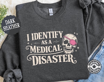 May include: Dark heather gray sweatshirt with the text "I IDENTIFY AS A MEDICAL DISASTER" and a skull wearing a pink eye patch. The sweatshirt has a crew neck and long sleeves. The words "DARK HEATHER" are printed on a black circle.