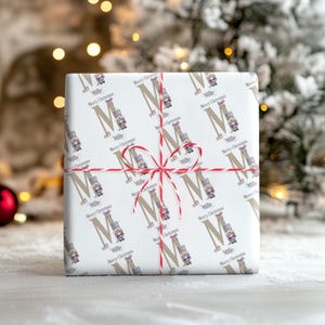 May include: A wrapped present with white paper. The paper has a repeating pattern of the letter "M", nutcrackers, and the text "Merry Christmas" and "Millie". It is tied with red and white twine.