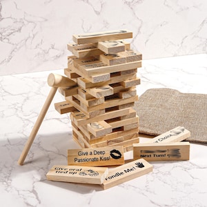 Pode incluir: Um jogo de empilhamento de blocos de madeira com um martelo, com blocos com instruções e ilustrações impressas. O jogo inclui uma torre de blocos, vários blocos soltos e um pequeno martelo. O texto nos blocos inclui frases como "Give a Deep Passionate Kiss!" e "Next Turn!"