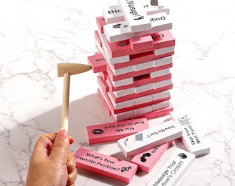 Puede incluir: Un juego de bloques de madera rosa y blanco con instrucciones impresas. Un pequeño martillo de madera se sostiene cerca de la torre. Algunos bloques están esparcidos. El texto en los bloques incluye "Foot Massage!" y "What's Your Favorite Position?"