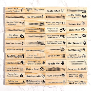 Puede incluir: Un conjunto de bloques de madera con varias frases e ilustraciones, diseñados para un juego. Los bloques presentan textos como "Taste Me!", "Get Naked!" y "Make Love to Me!", junto con gráficos relacionados. Los bloques están dispuestos sobre una superficie clara.