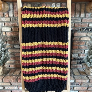 May include: A black, red, and gold striped knitted throw blanket draped over a wooden ladder.