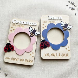 May include: Two wooden photo frames, one pink and one blue, each with a flower-shaped cutout. The pink frame says "Grandma" and the blue frame says "Mimi". Both have bee and ladybug embellishments and text that reads "Little Love Bugs".