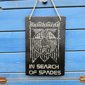 May include: Black slate sign with white text and graphic. The text reads "Hauntkind In Search Of Spades".