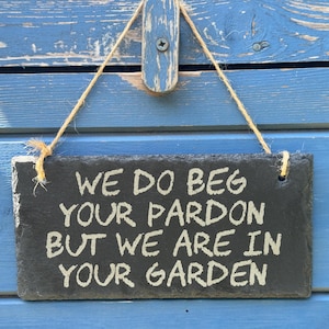 May include: A rectangular black slate sign featuring the engraved white text WE DO BEG YOUR PARDON BUT WE ARE IN YOUR GARDEN. The sign is suspended by a natural jute twine rope attached to a small wooden bracket at the top centre of the plaque.